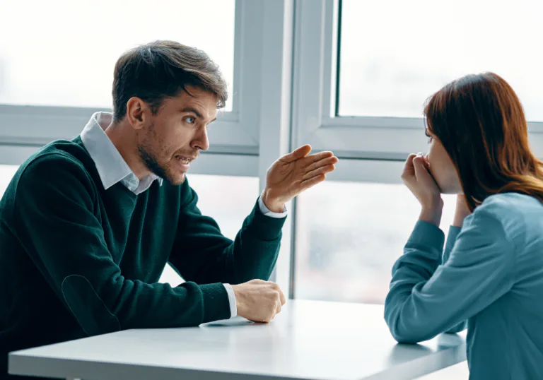 رجل يومئ بحماس أثناء شجار مع امرأة تجلس بهدوء إلى طاولة قرب نافذة.