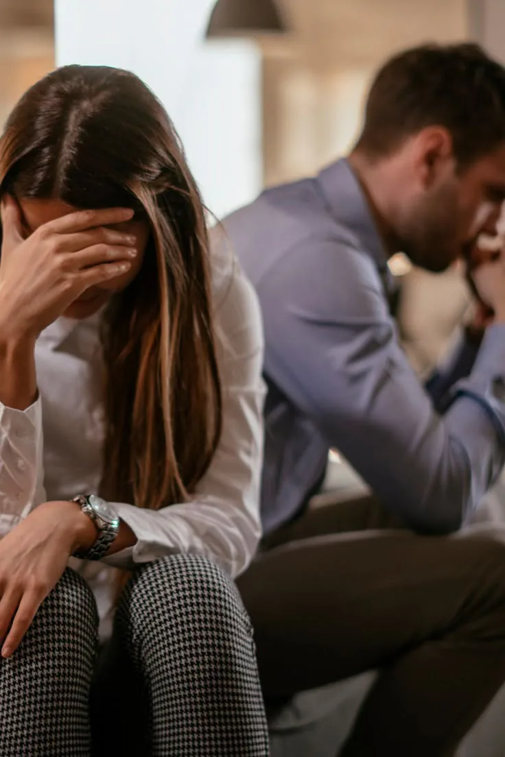 A distressed couple sitting apart after an argument, both looking upset and emotionally disconnected.
