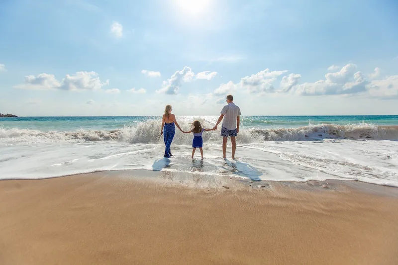Parents and child holding hands while standing in ocean waves on a bright beach