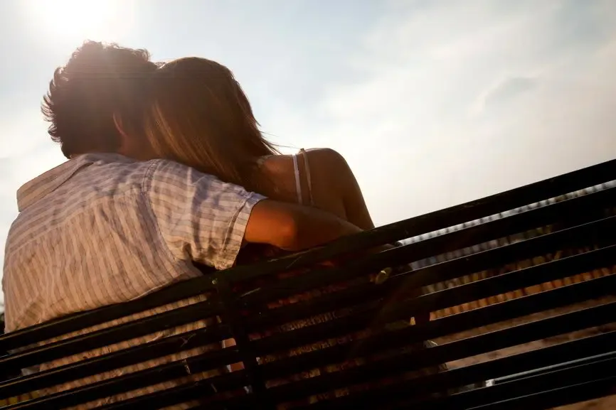 A couple sitting closely on a bench at sunset, embracing and sharing a tender, romantic moment.
