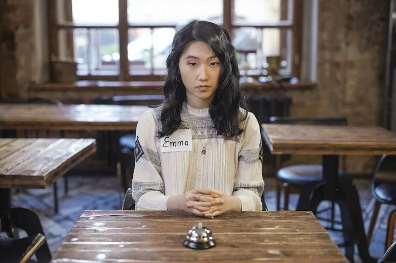 A young woman sitting alone at a wooden table with a thoughtful expression, suggesting emotional fatigue, reflection, or loneliness.