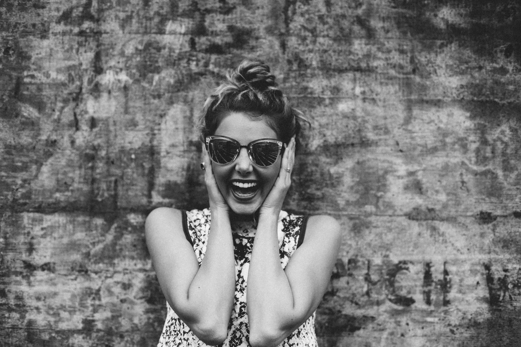 Smiling woman in sunglasses laughing with her hands near her face against a textured wall, expressing confidence and celebration.