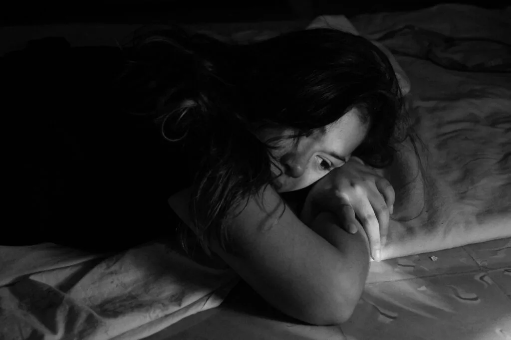 Woman lying on a bed in low light looking thoughtful and vulnerable, suggesting inner struggle, healing, and resilience.