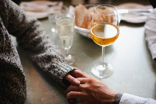 Two people holding hands across a table with drinks, symbolizing emotional support, intimacy, and comfort during graduation season.