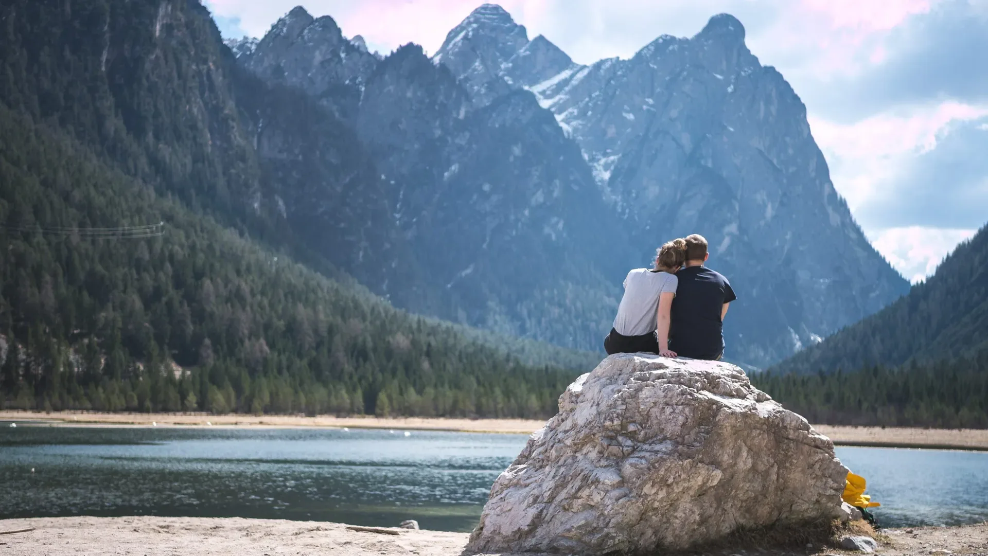 Couple sitting on a rock by a lake with mountains behind them, symbolizing shared journeys and achievement.